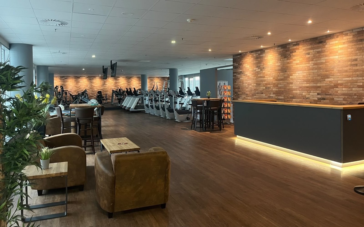 Lounge mit Ledersesseln und Bartischen neben dem beleuchteten Empfangs- und Wasserbar-Tresen im F4U Wiesbaden, dahinter die Cardio-Reihe an einer Backsteinwand